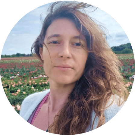A woman with long curly hair standing in a field of flowers. She is wearing a light sweater and appears to be outdoors on a cloudy day.